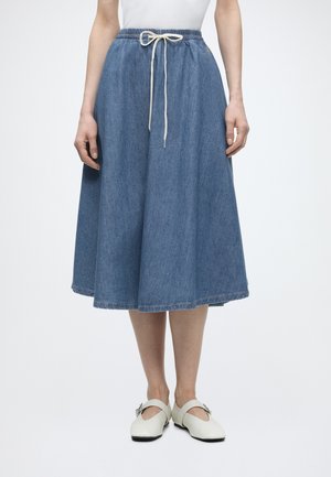 Kobieta w niebieskiej dżinsowej spódnicy do kolan z elastycznym pasem i białym sznurkiem, w połączeniu z białymi butami Mary Jane.