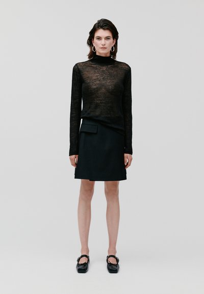Haut noir transparent à manches longues associé à une mini-jupe noire avec une poche avant. Le modèle porte des chaussures noires avec des boucles.