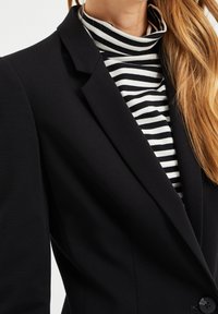 Svart kavaj med markerade axlar och en knappstängning över en svartvit randig polotröja, som visar kontrasterande texturer.