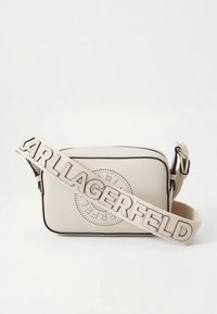 Beige Umhängetasche mit durchlöchertem Kreis-Design, schwarzem Rand und einem breiten Riemen mit groß geprägtem "LAGERFELD" Schriftzug.