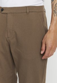 Brown cotton trousers with a textured weave. Features a front button closure, belt loops, and side pockets, shown at waist level.