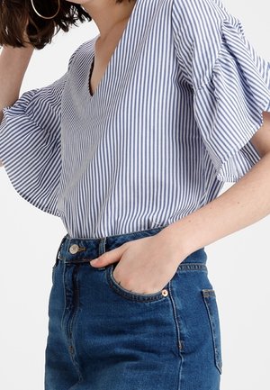 Femme portant un chemisier rayé bleu et blanc avec des manches à volants et un jean en denim bleu taille haute, main dans la poche.