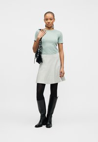 Γυναίκα με πλεξουδάκια στα μαλλιά, φοράει ανοιχτό πράσινο πουκάμισο, γκρι φούστα, μαύρο καλσόν, μαύρες μπότες μέχρι το γόνατο, κρατώντας μαύρη τσάντα ώμου.