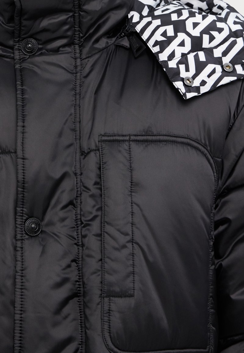 Chaqueta acolchada negra con acabado brillante, que presenta un gran bolsillo de parche y un cuello con un patrón en blanco y negro que contrasta.