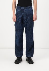 Persona vistiendo pantalones holgados de patrón azul marino, zapatos negros y una blusa sin mangas negra, de pie contra una pared blanca.