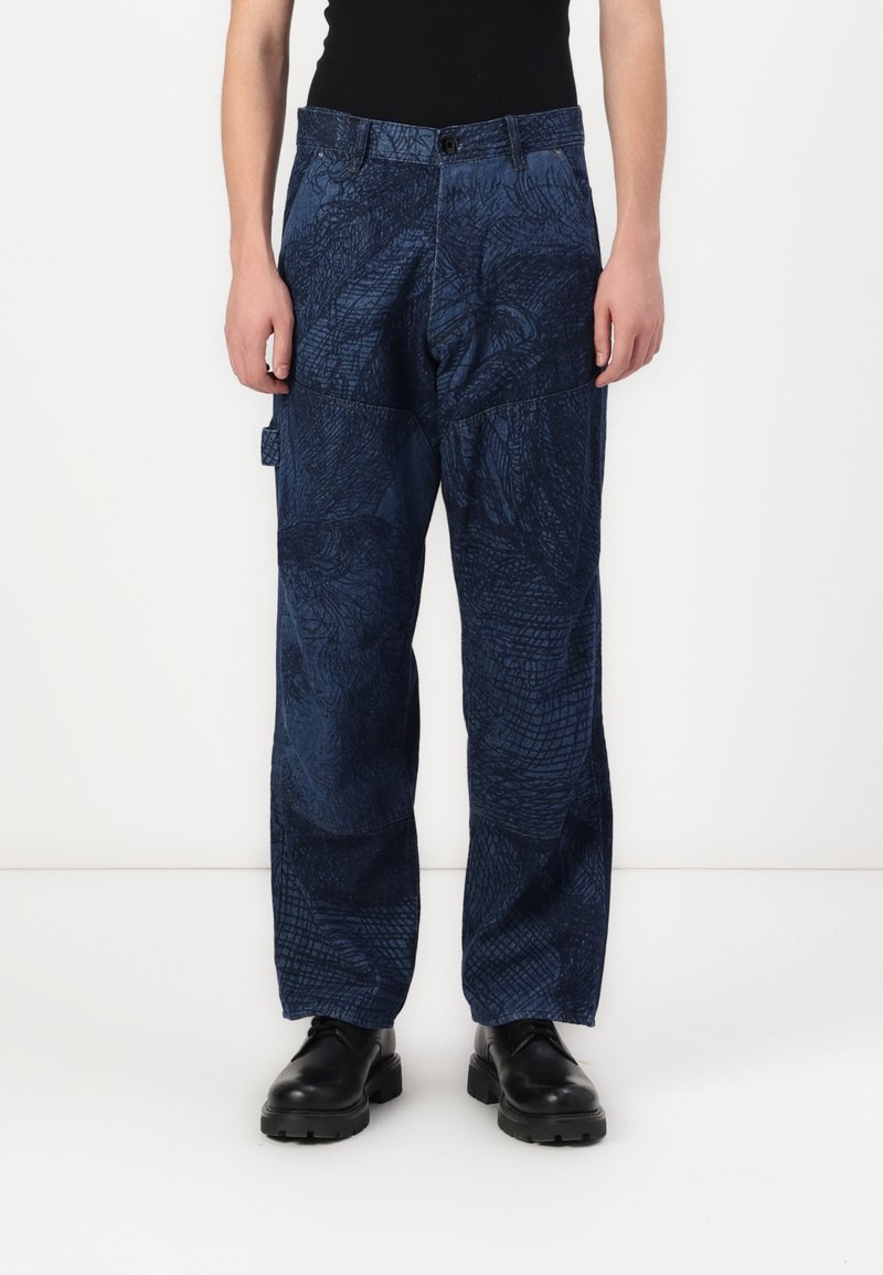 Persona vistiendo pantalones holgados de patrón azul marino, zapatos negros y una blusa sin mangas negra, de pie contra una pared blanca.