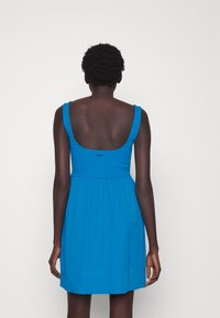 Blaues ärmelloses Baumwollkleid mit rückenfreiem Ausschnitt, tailliertem Bund und ausgestelltem Rock. Mit dezentem Branding oberhalb des hinteren Halsausschnitts.