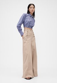 Sinisellä ja valkoisella raidallisella kauluspaidalla, yhdistettynä korkeavyötäröisiin beigen värisiin leveälahkeisiin housuihin, joissa nappikoristeet ja sivutaskut.