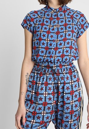 Kobieta ubrana w niebieski, wzorzysty top z krótkim rękawem oraz pasujące spodnie z elastyczną talią, ozdobione czerwonymi i białymi kwiatami oraz gwiazdkami, nosząca złoty naszyjnik.