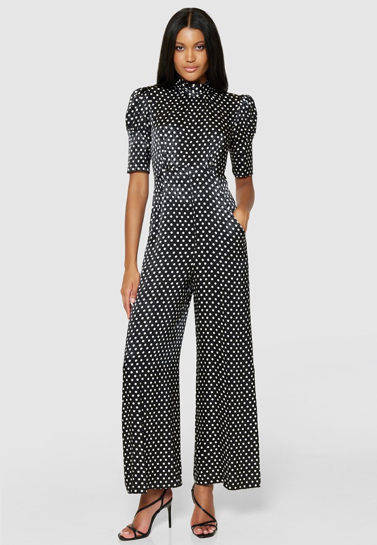 Czarny kombinezon w kropki z krótkimi bufkami, wysoką szyją, szerokimi nogawkami i kieszeniami. Gładki materiał o błyszczącym wykończeniu.