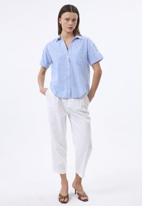 Camicia a righe blu e bianche a maniche corte con colletto e taschino frontale, abbinata a pantaloni di lino bianchi e sandali con tacco marroni.