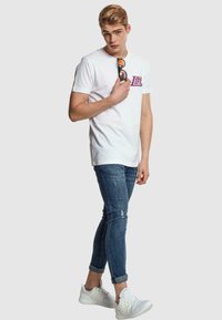 Tricou alb din bumbac cu un grafic purpuriu și mâneci scurte, asortat cu blugi albaștri distresați și teniși albi. Modelul poartă ochelari de soare.