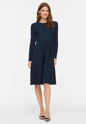 Femme debout portant une robe longue manches bleu marine, à étages, longueur genou, et des talons ouverts noirs sur un fond blanc uni.