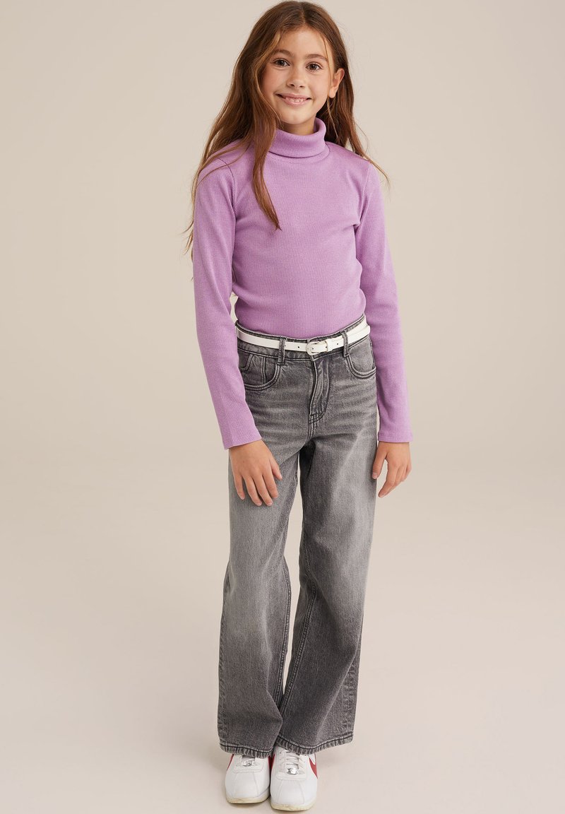 Pulover violet cu guler înalt, blugi gri evazați, curea albă, adidași albi cu accente roșii, fundal deschis. Copilul pozează cu mâinile relaxate.