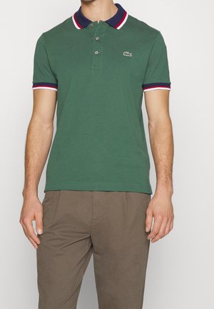 Groen poloshirt met een marineblauwe kraag en rood-wit gestreepte accenten op de mouwen. Voorzien van een knoopsluiting met drie knopen en het Lacoste-logo.