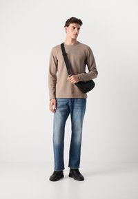 Model draagt een beige langslobig shirt, blauwe spijkerbroek en zwarte sneakers, terwijl ze een zwarte crossbodytas met een slank ontwerp en verstelbare schouderband vasthoudt.