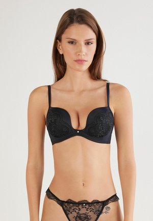 Soutien-gorge en dentelle noire avec décolleté plongeant, bonnets lisses et bretelles réglables. Tanga assorti en dentelle noire transparente, présentant des accents floraux et des bords festonnés.