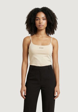 Vrouw met krullend haar, gekleed in een beige Guess-camisole en zwart broek met hoge taille, staand tegen een eenvoudige lichte achtergrond.