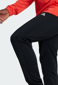 Pantaloni sportivi neri con un design elegante, caratterizzati da una trama sottile, un logo Adidas e una vestibilità flessibile. Giacca rossa visibile nell'inquadratura.