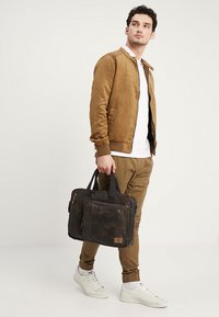 Braune Wildleder-Bomberjacke, weißes T-Shirt und khakifarbene Jogginghose. Hält eine dunkelbraune Lederhandtasche mit einer vorderen Tasche und zwei Griffen.