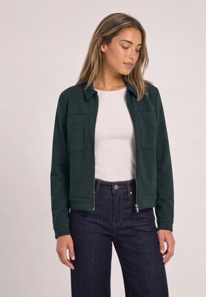 Une femme porte une veste vert foncé zippée sur le devant, avec des poches poitrine, par-dessus un haut blanc et un jean foncé.