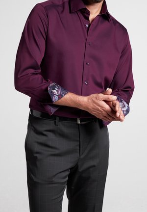 Uomo che indossa una camicia viola con bottoni e motivo floreale sui polsini arrotolati, abbinata a pantaloni da abito scuri a quadri e una cintura nera.