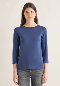 Femme aux longs cheveux bruns portant une chemise bleue à manches longues et un jean gris, debout devant un fond clair uni.