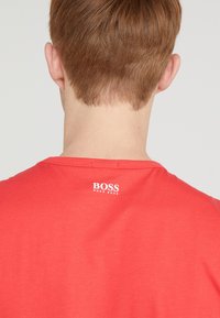 Červené bavlněné tričko s kulatým výstřihem a krátkým rukávem, na zadní části krku je bílá loga "BOSS". Hladká textura.