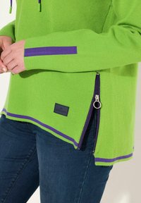 Golf verde con accenti viola, caratterizzato da una zip laterale e un tessuto testurizzato. Etichette visibili sul davanti e sulla cucitura laterale.