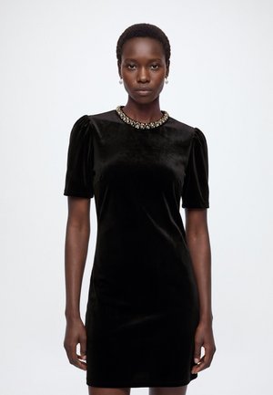 Femme portant une robe courte en velours noir avec des épaules bouffantes et un décolleté orné de bijoux, debout devant un fond blanc uni.