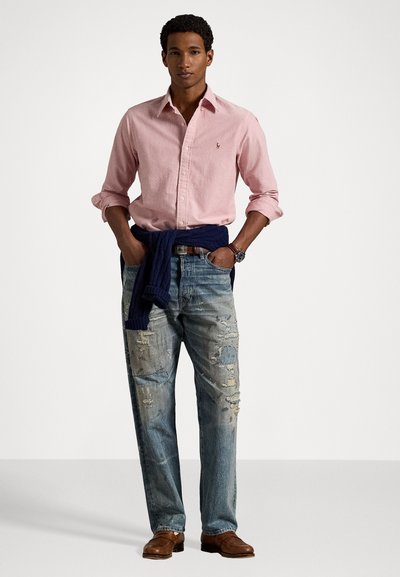 Ljusrosa skjorta, blå tröja knuten i midjan, slitna blå jeans, bruna skor och silverfärgad armbandsur.