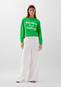 Felpa verde con testo bianco che recita "Il paradiso è una sensazione", abbinata a pantaloni bianchi larghi. Tessuto elegante e vestibilità comoda.