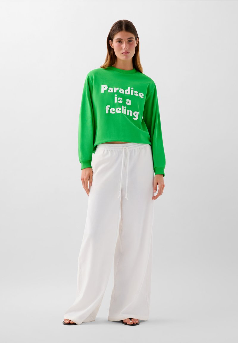 Grüner Pullover mit weißem Text "Paradise is a feeling" kombiniert mit lockeren weißen Hosen. Schlichte Stoffe und entspannter Schnitt.
