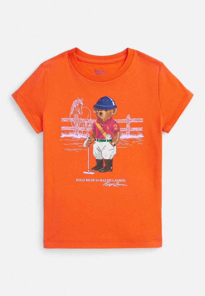 Spoža oranža T-krekls ar lāča grafiku polo gear, turot āmuru, stāvot priekšā zirgam un žoga kontūrai.