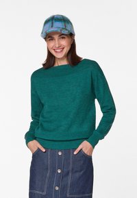 Grüner Strickpullover mit einem Boot-Ausschnitt, getragen über einem Jeansrock mit vorderen Knöpfen. Eine blau-karierte Mütze rundet das Outfit ab.
