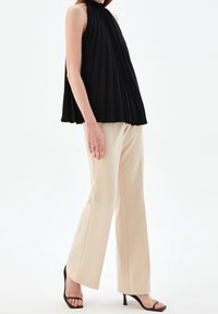 Kvinna som bär en ärmlös svart plisserad topp, beigefärgade vida byxor och svarta klacksandaler, stående framför en vit bakgrund.
