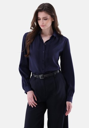 Marineblaue Seidenbluse mit Knopfverschluss, langen Ärmeln und subtilen Falten. Kombiniert mit hochgeschnittenen schwarzen Hosen und einem strukturierten Gürtel.