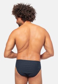 Uomo con i capelli ricci, di spalle rivolto verso la telecamera, che indossa slip da bagno blu scuro con sottili strisce bianche verticali.