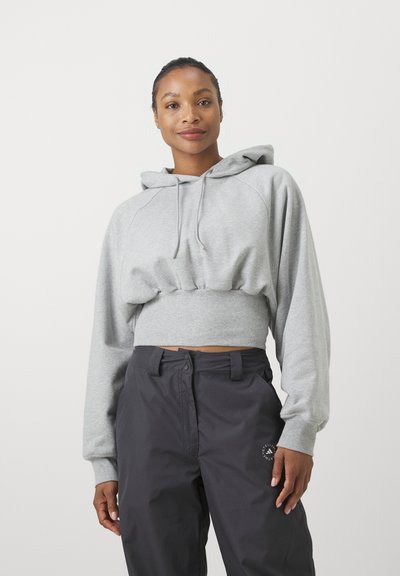 Mujer con una sudadera corta con capucha de color gris claro y pantalones negros de cintura alta con un pequeño logo redondo en el muslo izquierdo, de pie frente a un fondo blanco.