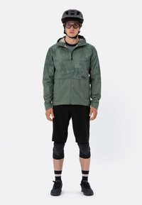 Lättviktig grön vattentät jacka med huva och svarta shorts. Utrustad med vadderade knäskydd, sneakers och en svart hjälm med glasögon.