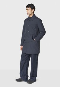 Navyblauer, knielanger Mantel mit Knopfleiste, Seitentaschen und strukturiertem Kragen. getragen über locker sitzenden, dunklen Denim-Hosen.
