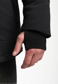 Iš juodos striukės rankovės, kurioje yra angos nykščiui, išlenda ranka prieš baltą foną.