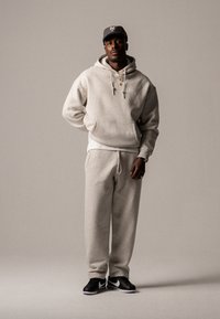 Hellgrauer Pullover und passende Jogginghose mit einer Fronttasche und Kordelzug. Schwarze Sneaker mit weißen Akzenten. Flachschirmmütze.