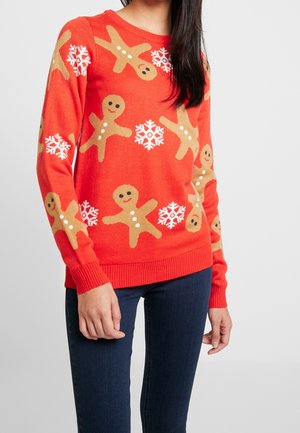 Femme portant un pull rouge orné de motifs de bonhommes en pain d'épice souriants et de flocons de neige blancs, assorti à un jean bleu foncé.