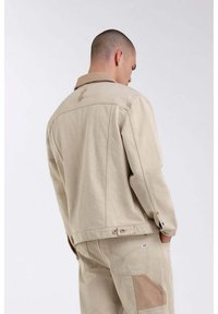 Giacca di cotone beige con colletto classico, bottoni ai polsini e dettagli di cucitura discreti. Si abbina a pantaloni cargo beige.