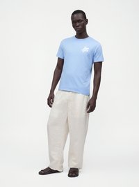 Homme portant une chemise bleu clair à manches courtes avec un motif de tortue blanche, un pantalon blanc ample et des sandales foncées, debout devant un fond blanc uni.