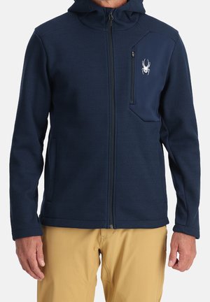 Mand iført navyblå lynlåshættejakke med edderkop-logo på brystlommen og beige bukser, står mod en ensfarvet baggrund.