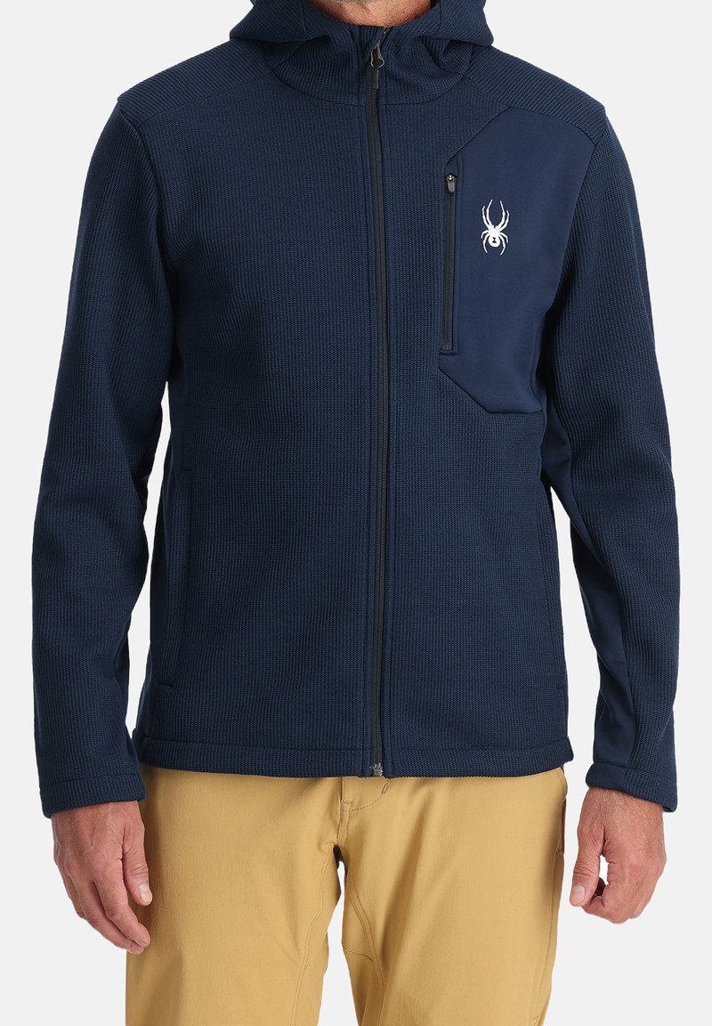Homme portant un sweat à capuche bleu marine zippé avec un logo d’araignée sur la poche poitrine et un pantalon beige, debout devant un fond uni.