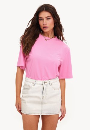 Femme aux longs cheveux bruns portant un haut rose à manches courtes ample rentré dans une minijupe en jean clair, debout devant un fond blanc.