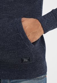 Marinblå stickad tröja med ett strukturerat mönster, som har en framficka och ribbade muddar. En liten etikett visar "PIER-ONE"-logotypen.